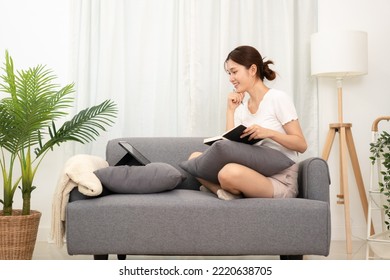 Young Asian Woman Looking Data On Tablet And Writing New Idea Of Project In Notebook While Wearing Casual Clothes To Sitting And Working On Comfort Sofa In Living Room At Home.