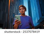 Young Asian woman looking at her mobile phone while standing on the street at night, illuminated by city lights, focused on screen, possibly texting or browsing social media, urban evening atmosphere