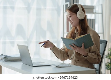 Young Asian woman looks happy wearing headphones Watch the webinar Listen to online courses Communicate with video conferencing and learn languages Laughing at teacher's lesson on laptop at desk. - Powered by Shutterstock