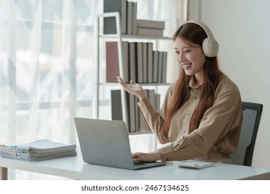 Young Asian woman looks happy wearing headphones Watch the webinar Listen to online courses Communicate with video conferencing and learn languages Laughing at teacher's lesson on laptop at desk. - Powered by Shutterstock