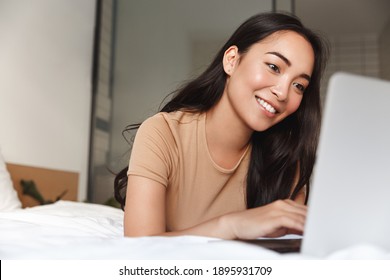 Young asian woman lying in bed at home and using laptop. Girl smiling happy while looking at computer.
