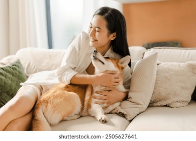 Young asian woman is lying on a comfortable sofa in a bright living room, gently hugging her cute pembroke welsh corgi dog - Powered by Shutterstock