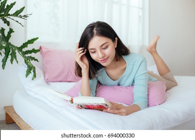 Young Asian Woman Lying On Bed, Reading Book