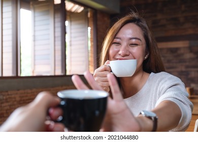 A Young Asian Woman Making Fun By Making Hand Sign To Refuse Clinking Coffee Mug With Her Friend