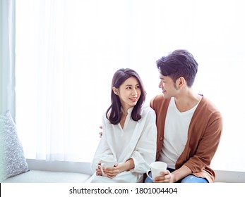 Young Asian Woman And Man At Home With Cup Of Coffee In Hands At Home During COVID-19