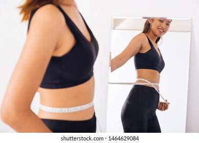 Young Asian woman measuring waist with tape in front of a mirror. Smiling sporty girl in sport bra looking at reflection. - Shutterstock ID 1464629084