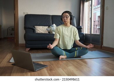 Young asian woman meditating following a meditation online class on her laptop at home. Easy pose (Sukhasana).