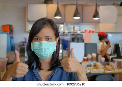 Young Asian Woman Or Owner Store Wearing Medical Face Mask During Covid19 Outbreak In Coffee Shop To Protect From Coronavirus, Care For Health And Safety, Showing Good Gesture Sign.