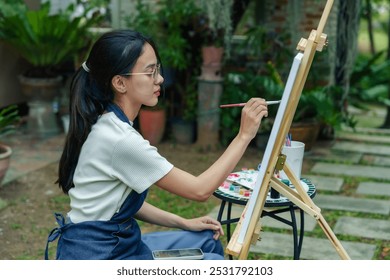 Young Asian woman painting on canvas in garden setting. Focused expression while creating artwork outdoors. Surrounded by greenery, sitting near easel with art supplies. Relaxed, creative atmosphere. - Powered by Shutterstock