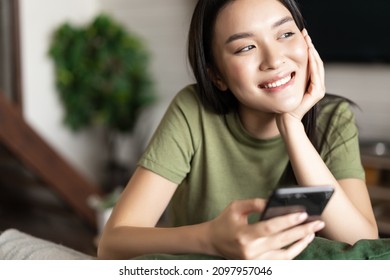 Young Asian Woman Placing An Order, Waiting For Delivery On Mobile Phone App, Sitting At Home And Looking Outside Window