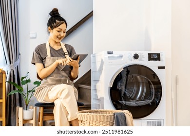 Young asian woman are playing with digital tablet in home sitting near the washing machine, while waiting for the washing machine to finish working - Powered by Shutterstock