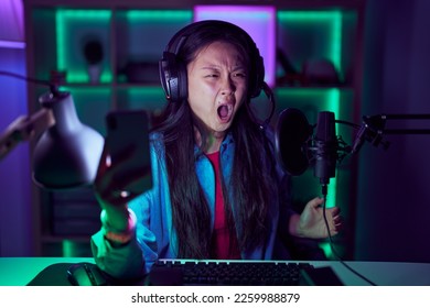 Young asian woman playing video games with smartphone angry and mad screaming frustrated and furious, shouting with anger. rage and aggressive concept. 