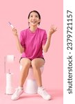 Young Asian woman with pregnancy test sitting on toilet bowl against pink background