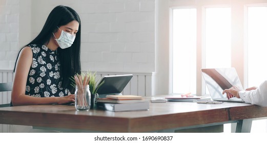 Young Asian Woman With Protective Mask Woking With Her Friend While Sitting At Home Office. Businesswoman Wearing Mask To Protect Her Health. Working From Home Concept.