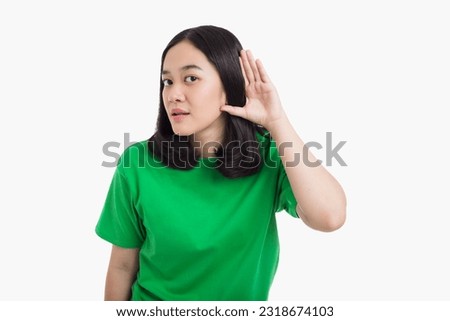 Young Asian woman puts a hand over ear listening conversation or something wearing green t-shirt standing over white background