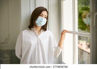 Young Asian Woman In Quarantine Wearing Mask Standing By Window Looking Sad And Depressed