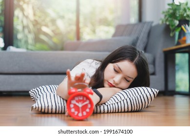 Young Asian Woman Reaching Over To Turning Off Alarm Clock While Lying On The Floor In Living Room At Home.