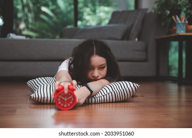 Young Asian Woman Reaching Over To Turning Off Alarm Clock While Lying On The Floor In Living Room At Home.