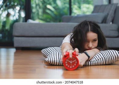 Young Asian Woman Reaching Over To Turning Off Alarm Clock While Lying On The Floor In Living Room At Home.