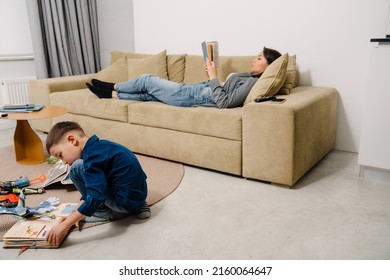 Young Asian Woman Reading Book On Sofa While Her Little Son Playing With Toys On Floor At Home