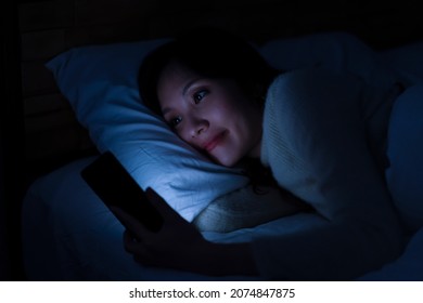 Young Asian woman reading the text message on a smartphone in bed at night with smiling face before falling sleep, going to bed late.  - Powered by Shutterstock