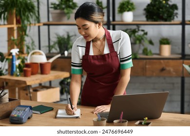 A young Asian woman in a red apron works at her plant store, taking inventory or managing orders. - Powered by Shutterstock