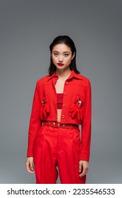 young asian woman in red jacket decorated with brooches and gloves looking at camera isolated on grey - Shutterstock ID 2235546533