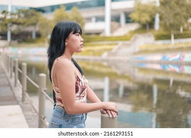 A Young Asian Woman Reflects On Old Memories Or Pondering On Her Relationship And Career. Experiencing Quarter Life Crisis. At A Walkway By A Public Promenade.