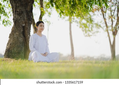 Young asian woman relaxes wearing white dress meditation at green grass fields, Thai woman Buddhism meditation in Buddhist holy day