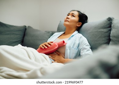 Young Asian Woman Relieves Stomach Pain With Hot Water Bottle While Relaxing On Sofa At Home.