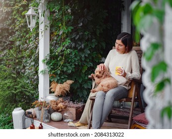 Young asian woman resting outdoors with cup of coffee on porch of country house, chilling outside with poodle dog . autumn lifestyle, leisure free time concept. Copy space