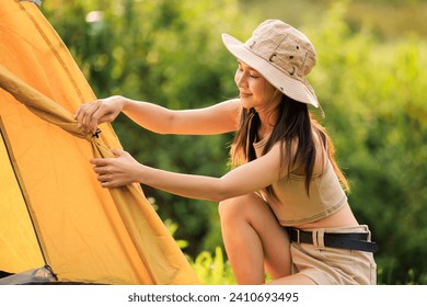 young asian woman in safari dress setting up tent by herself, at natural park campground,