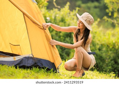 young asian woman in safari dress setting up tent by herself, at natural park campground,