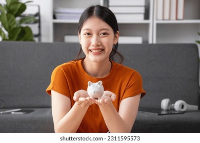 Young Asian woman saving for retirement Saving money through a piggy bank and taking notes on notebook, savings concept. - Shutterstock ID 2311595573
