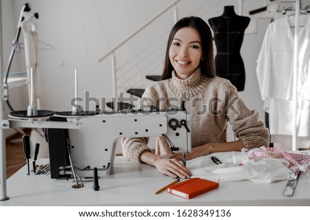 Young asian woman seamstress at her sewing machine smiling and looking at camera. Sewing, design work, tailoring studio, tailor, designer clothes, manufactory, in the process of creative development