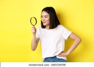 Young Asian Woman Searching For Something, Looking Left Through Magnifying Glass And Smiling Pleased, Investigating, Standing Over Yellow Background