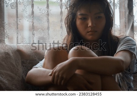 A young Asian woman with short hair and a white t-shirt sits with her arms crossed and her knees crossed, looking lost on the sofa by the window.