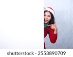 young asian woman shouting happily to camera hand pointing to right, wearing red christmas sweater, red glasses, santa hat and scarf, behind blank space board isolated on white background