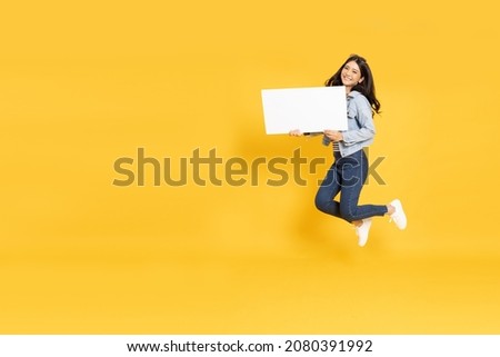 Young Asian woman showing and holding blank white billboard and jumping isolated on yellow background
