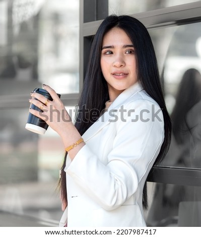 Young Asian woman sits at cafe window drinking a cup of tea or coffee, portrait. Beautiful girl leisure relaxation in coffee shop with hot drink beverage. Attractive lady takes a refreshment break.