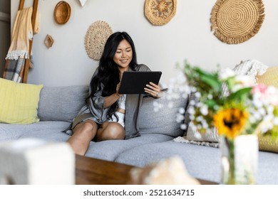 A young Asian woman sits comfortably on a sofa, using a tablet. Cozy living room with a warm atmosphere for relaxation and enjoyment. Asian woman using a digital tablet, social media content creator. - Powered by Shutterstock