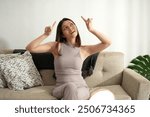 A young Asian woman sits on a cozy sofa in a bright living room, pointing upwards with both hands. She appears curious and thoughtful, enjoying a relaxed moment at home during the day.