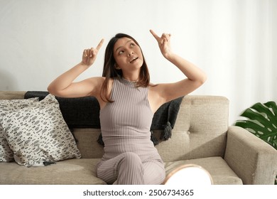 A young Asian woman sits on a cozy sofa in a bright living room, pointing upwards with both hands. She appears curious and thoughtful, enjoying a relaxed moment at home during the day. - Powered by Shutterstock