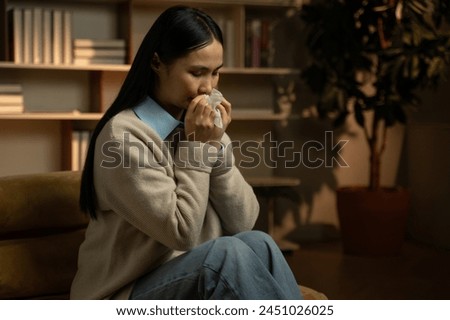 A young Asian woman is sitting comfortably in an indoor setting, possibly her home, and appears to be unwell as she is blowing her nose with a tissue. She seems to be experiencing cold symptoms and is