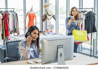 Young Asian Woman Sitting In Front Of Computer In A Clothing Shop Talking On Mobile Phone. A Customer Also Holding Mobile Phone In The Background.