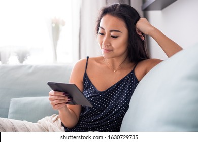 Young asian woman sitting on sofa using reader tablet and reading a ebook at home