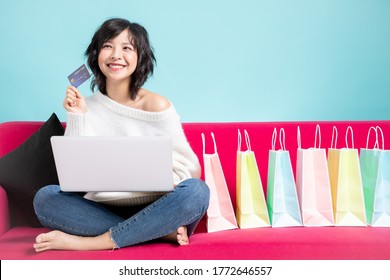 Young Asian Woman Sitting On Sofa With Laptop Computer And  A Credit Card In Her Hand Showing Plenty Of Colorful Shopping Bags, Isolated On Light Blue Background.