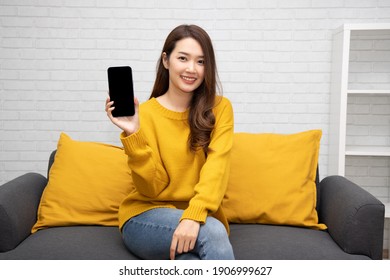 Young Asian Woman Sitting On Sofa And Showing Present Mobile Phone Application At Home