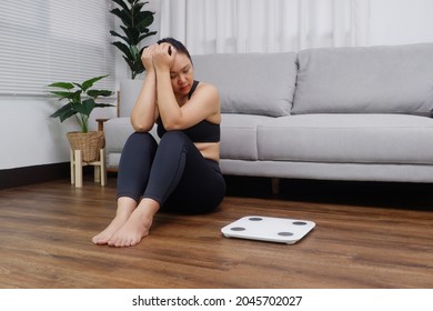 Young Asian woman sitting on the floor looking at the weight scale worried and depressed in dieting and eating disorder concept