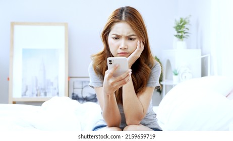 Young Asian woman sitting on bed using smartphone with bad mood and frowning face in bedroom. - Shutterstock ID 2159659719